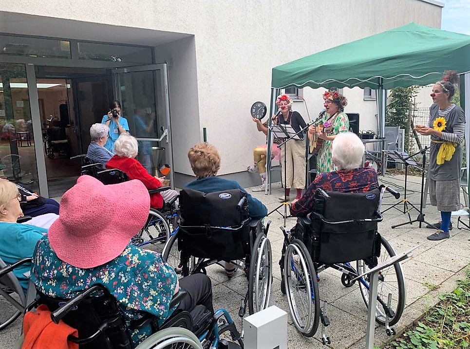 Un groupe de personnes âgées en fauteuil roulant observe trois artistes vêtus de costumes et jouant des instruments de musique sous un auvent vert, à l'extérieur d'un bâtiment. Un photographe se tient à l'arrière-plan, près d'une porte vitrée ouverte.
