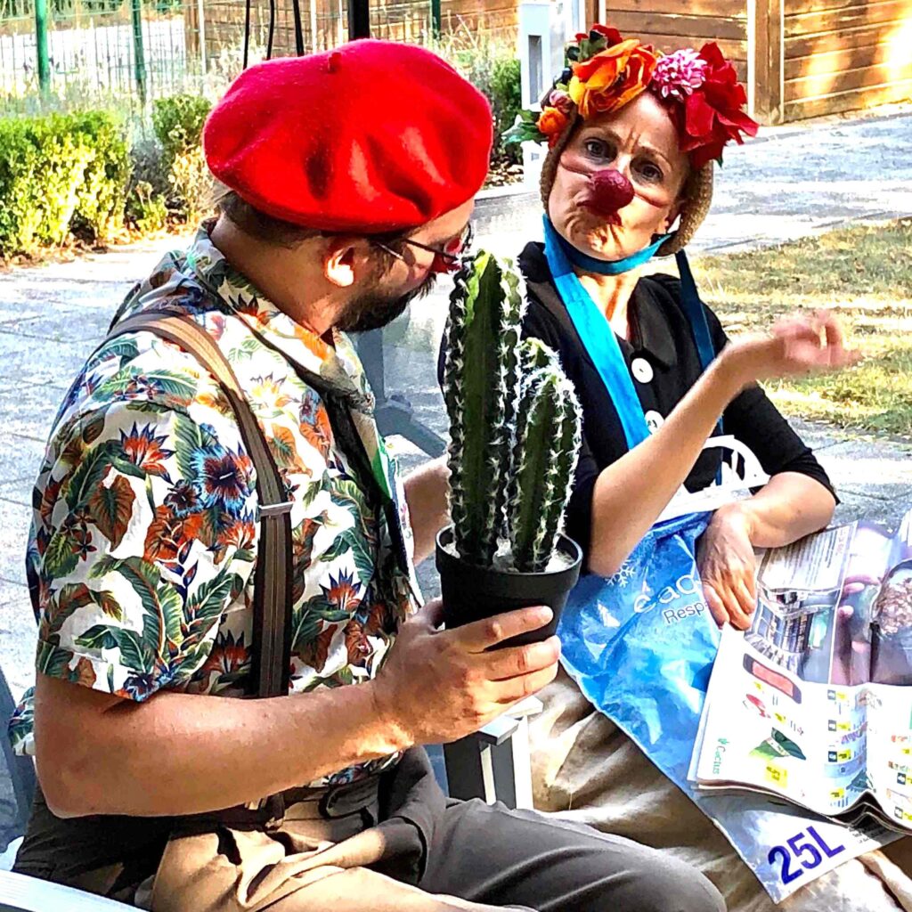 Deux personnes déguisées en clowns sont assises à l'extérieur ; l'une tient un cactus en pot et l'autre fait des gestes théâtraux, coiffée d'un nez rouge et d'un bandeau à fleurs. Toutes deux portent des costumes colorés et semblent engagées dans une conversation enjouée.