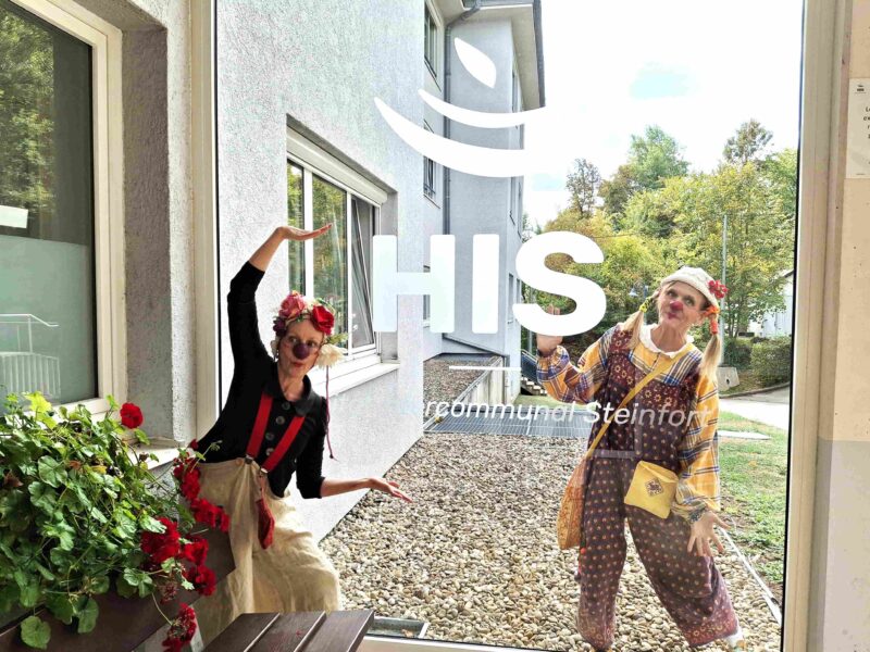 Deux personnes vêtues de costumes colorés prennent la pose et sourient derrière une vitrine sur laquelle sont inscrits « HIS » et « Communauté Steinfotz ». Des fleurs rouges sont disposées sur une table au premier plan, tandis qu'un bâtiment et des arbres sont visibles à l'extérieur.