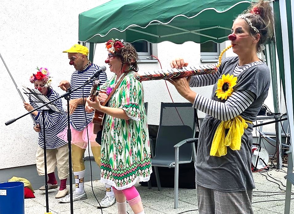Quatre personnes déguisées en clowns espiègles jouent de la musique à l'extérieur, sous un auvent vert. Elles portent des costumes colorés, des bandeaux fleuris et des nez rouges. L'une d'elles joue d'un instrument amusant, une autre de la guitare, et deux chantent au micro.