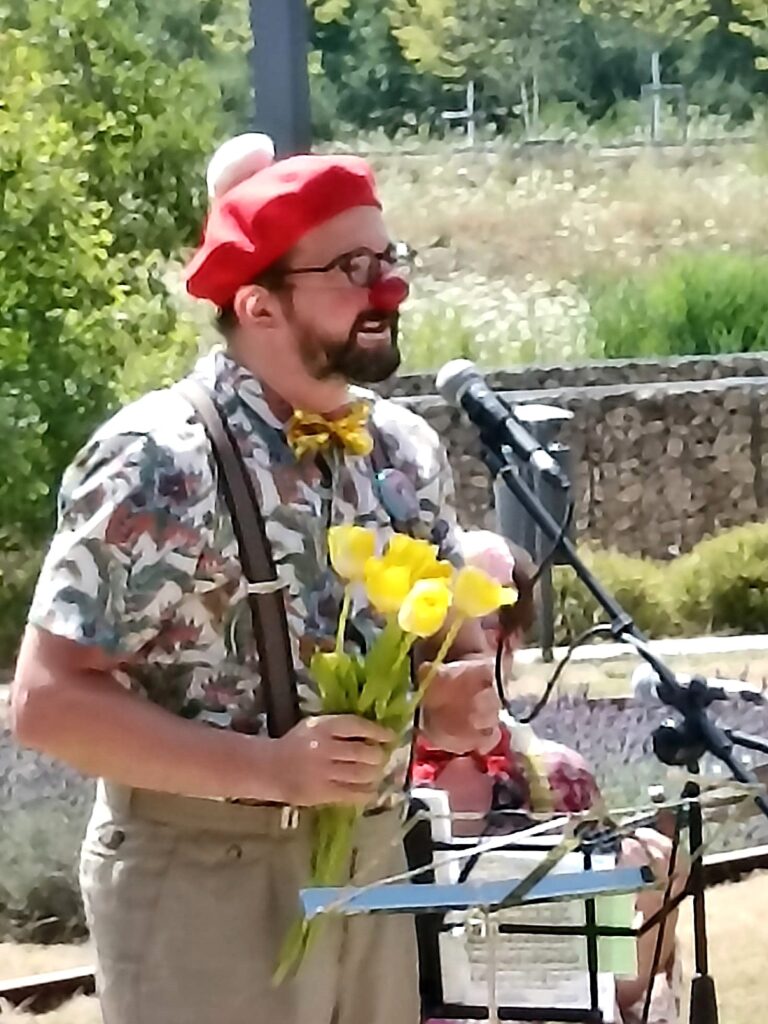Un homme déguisé en clown, coiffé d'un chapeau rouge et portant un nœud papillon, tient des fleurs jaunes et parle ou chante dans un micro à l'extérieur.