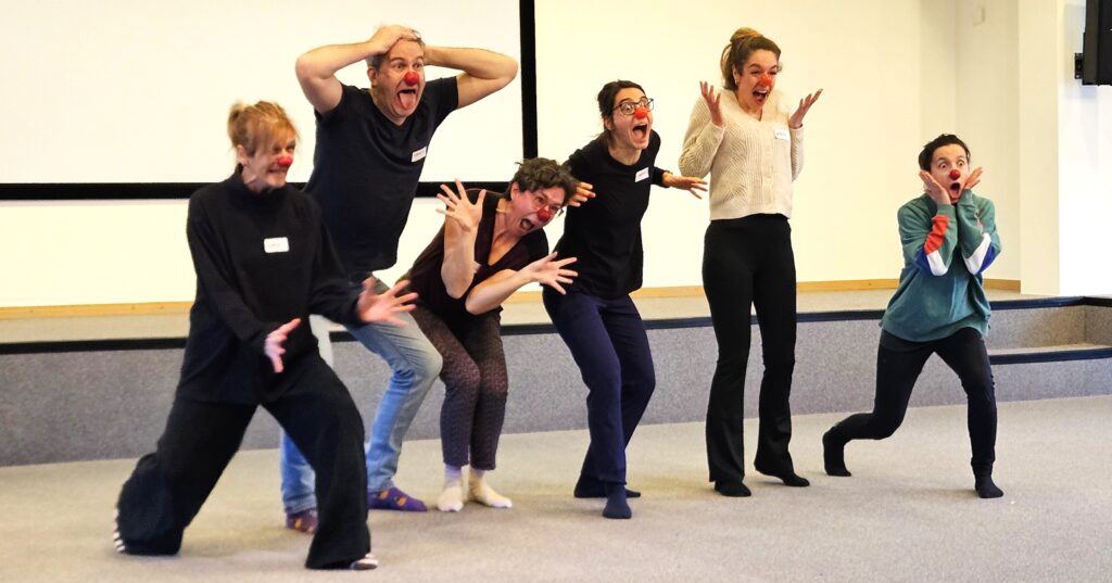 Six adultes portant des nez rouges de clown sont alignés à l'intérieur ; ils font des mimiques exagérées et des gestes théâtraux, comme s'ils jouaient la comédie ou feignaient la surprise.