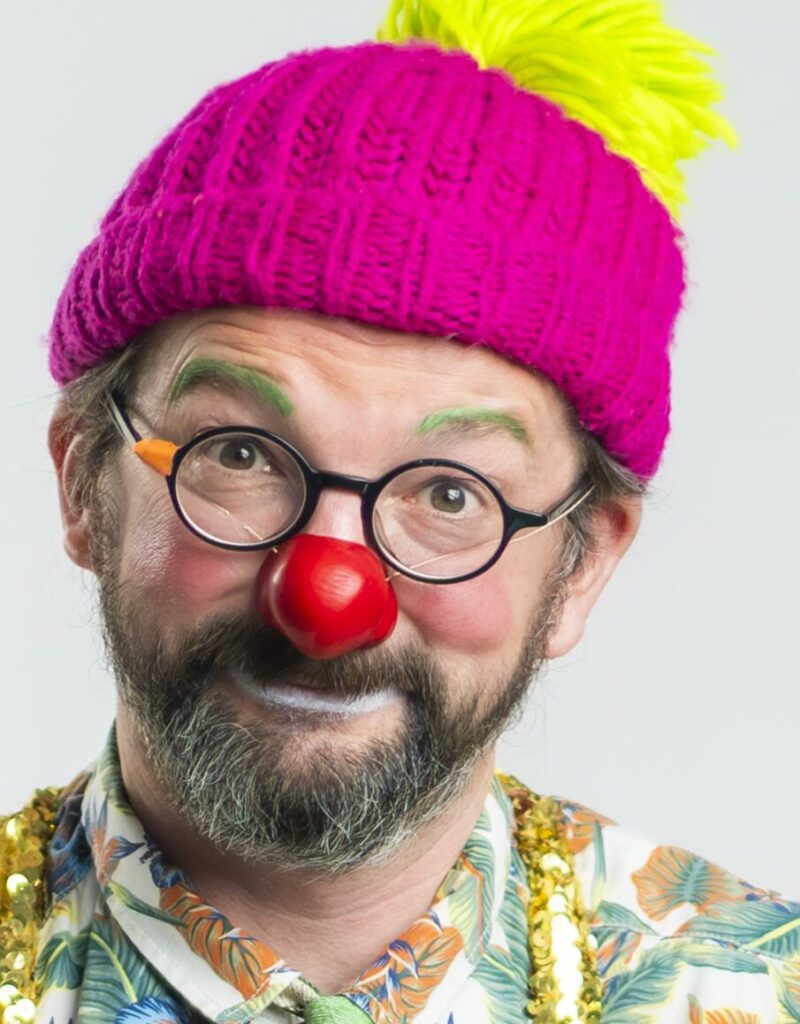 Un homme déguisé en clown, coiffé d'un chapeau rose vif orné d'un pompon jaune, portant des lunettes rondes, un nez rouge, des sourcils verts et une chemise colorée, sourit à la caméra.