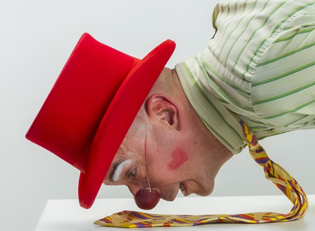 Un clown au nez et au chapeau rouges, le visage peint, vêtu d'une chemise rayée et d'une cravate colorée, se penche sur le côté, la joue appuyée contre une surface blanche.