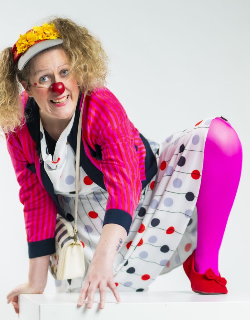 Un clown souriant, aux cheveux bouclés, avec un nez rouge, un bandeau jaune, une veste à rayures roses, une robe à pois, des collants rose vif et des chaussures rouges, est accroupi de manière enjouée sur une surface blanche, devant un fond blanc.