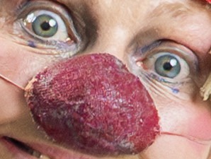 Gros plan sur une personne portant un grand nez de clown rouge, avec de grands yeux verts bien ouverts, du maquillage autour des yeux et des ficelles visibles qui maintiennent le nez en place.