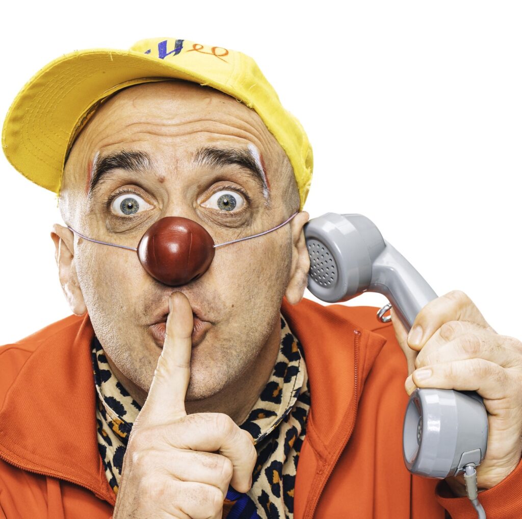Une personne déguisée en clown, avec un nez rouge et une casquette jaune, tient un téléphone contre son oreille et fait le geste « chut » en portant un doigt à ses lèvres.