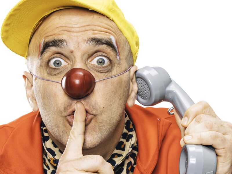 Une personne déguisée en clown, avec un nez rouge et une casquette jaune, tient un téléphone contre son oreille et fait le geste « chut » en portant un doigt à ses lèvres.