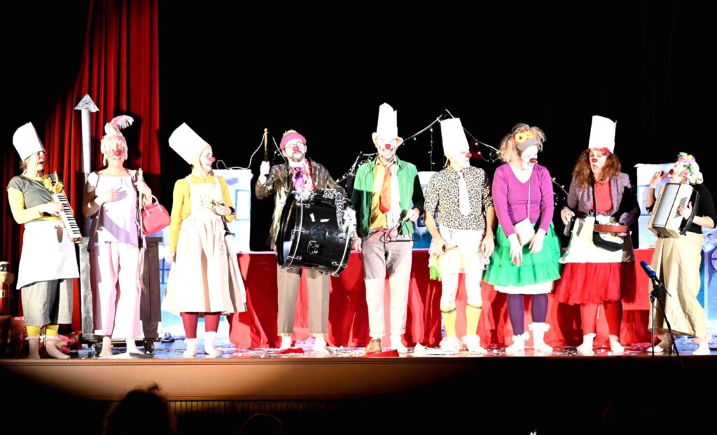 Neuf personnes vêtues de costumes de clowns colorés et coiffées de toques de chef se tiennent sur une scène devant un rideau rouge, jouant de leurs instruments de musique et souriant.