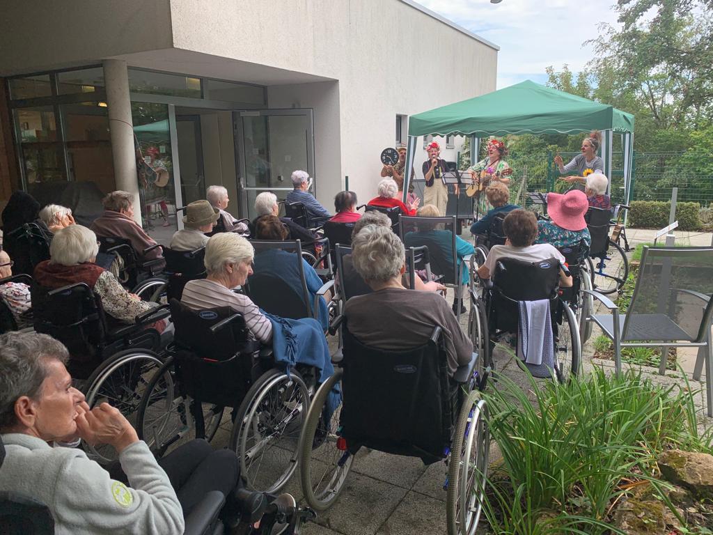 Un groupe de personnes âgées en fauteuil roulant est assis à l'extérieur, face à des musiciens qui se produisent sous un auvent vert. Le lieu semble être un établissement de soins doté d'un jardin et d'une architecture moderne.
