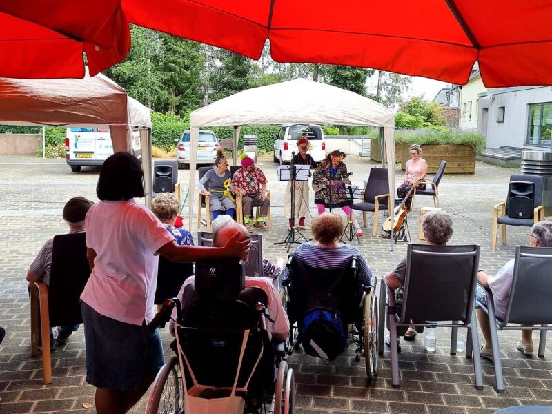 Un petit concert en plein air où des musiciens se produisent sous un auvent devant un public, dont certains en fauteuil roulant, assis sous des parasols rouges dans une cour pavée.